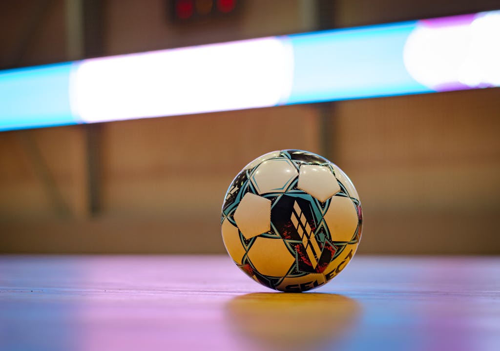 Close-up of a soccer ball on a gym floor with blurred background lights.